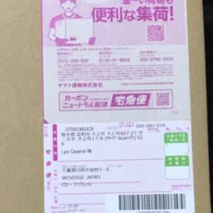 Pink Japanese parcel label on a cardboard box, featuring a cartoon courier, a QR code, a barcode, and the recipient's address in Japanese.