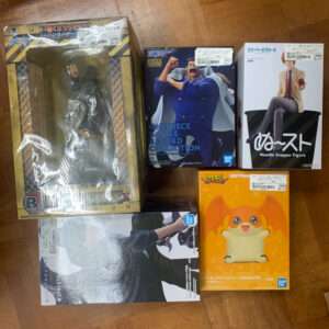 Six boxed figures on a wooden floor, some in plastic wrap, including a One Piece Battle Record Collection box and an orange bat-like creature figure.