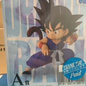 Dragon Ball action figure of Goku in a blue gi with a gray sash, crouching in a fighting pose with a brown tail visible, on packaging with large blue letters in the background.