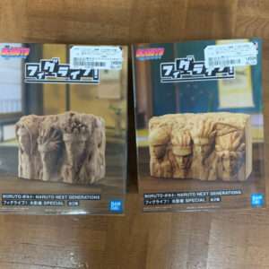 Two boxed Boruto/Naruto Next Generations figures on a wooden floor, each box showing a carved wooden block with multiple faces and characters.