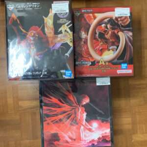 Three boxed anime figures on a wooden floor: Evangelion figure left, One Piece figure right, and a third red-themed artwork box below.