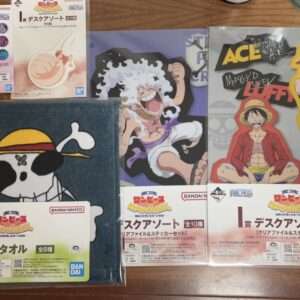 Collection of One Piece merchandise on a wooden table: a Skull pirate towel, two character posters, and sticker/card sets featuring Luffy and Ace with Bandai Namco packaging.