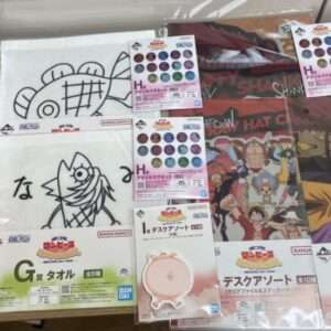 Display of One Piece merch on a wooden surface: a boxed figure on the left, white towels with drawn character designs in the center, and large colorful crew posters/cards on the right.