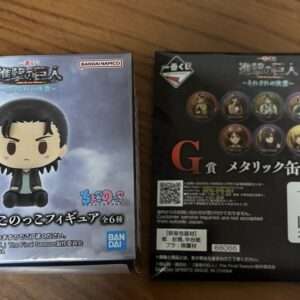 Two boxed chibi-style anime figures on a wooden surface; left box features a small figure with black hair and a blue jacket labeled H prize, Bandai Namco branding visible. Right box shows eight circular metal can badges with character portraits and a QR code on the back, also Bandai Namco branding.