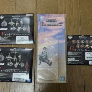Boxed anime merchandise on a wooden floor: two black F acrylic charm sets and a G badge set plus a blue E standee box with a seated figure in plastic casing.
