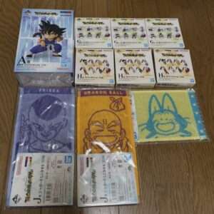 Assorted Dragon Ball merchandise on a wooden floor: a large Goku action figure box, several small figure boxes, and three character towels featuring Frieza, Krillin, and a blue cartoon character.
