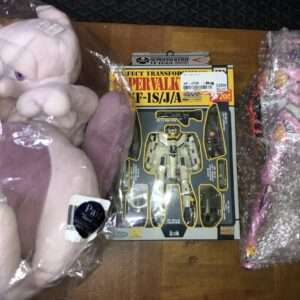 Pink plush toys in plastic bags surround a boxed robot action figure on a wooden floor, seen from above.