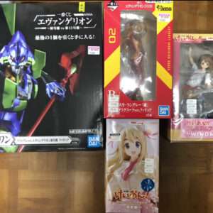 Large Evangelion unit figure box on the left with bold blue and green mecha art, brand Bandai Megahouse, on a wooden floor. To the right are three smaller boxed figures: a red-and-yellow box featuring Asuka Langley figure visible through a window, a pink box with a Windmill anime figure, and a smaller box showing a blonde anime girl in a white outfit.