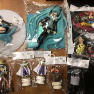 Top-down view of a collection of packaged anime figurines laid out on a wooden surface, including multiple Hatsune Miku figures and other characters with price tags.
