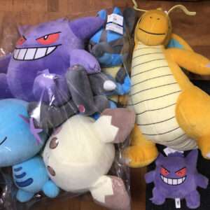 A pile of colorful plush toys in a cardboard box, including a purple monster with red eyes, a light blue sea creature plush, and a large yellow dragon with a striped belly and horns in the corner.