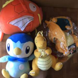 Several large plush toys on a wooden floor: an orange fish-like character, a blue penguin, and a yellow dragon, with one in clear packaging.