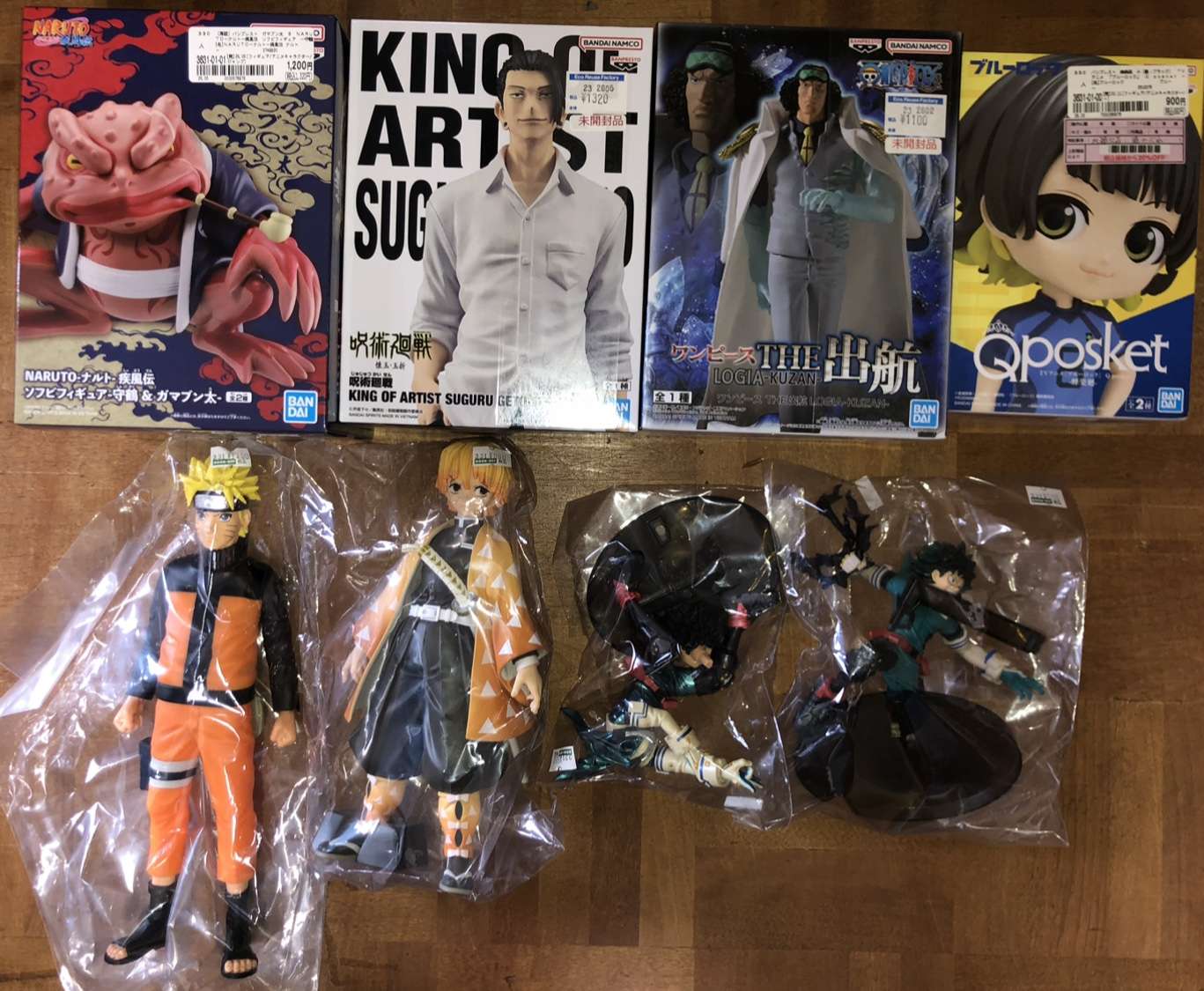 Screenshot - Articulos Japan (post Box Japan) Four boxed anime figure sets arranged on a wooden floor (left to right: a Naruto creature figure, King of Artist Suguru, Logia Kuzan from One Piece, and Qposket character), with four loose figures still in plastic bags laid out below. Articulos Japan (Post Box Japan)