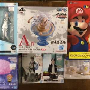 Boxed anime figures on a wooden floor: Evangelion Rei (left), One Piece statue (center), Super Mario box (right), plus smaller boxes below.