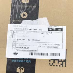 Close-up of a cardboard parcel with a white shipping label, showing a barcode, date 04/28, and Amazon.co.jp contact details.