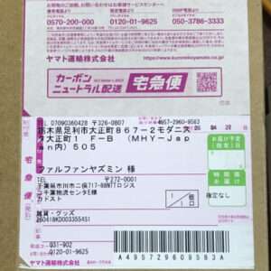 Parcel delivery label on a cardboard box with a barcode, tracking numbers, and recipient/sender fields visible under a pink-marked area (Japanese shipping label).  Articulos Japan (Post Box Japan)