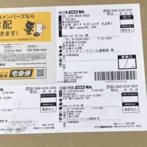 Sheet of Japanese shipping labels with barcodes on a brown surface; left side shows a yellow ad with a black cat mascot.  Articulos Japan (Post Box Japan)