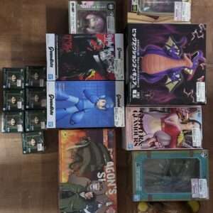 Assortment of boxed anime and video game figures on a wooden floor, including a blue Mega Man figure and a large purple dragon box, with a bear plush in plastic on the right corner and smaller character boxes on the left edge.