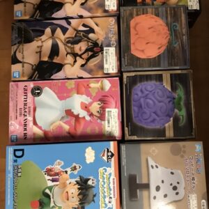Row of boxed anime figures and collectibles arranged on a wooden floor, including two identical boxes of a female character in a revealing outfit, a pink-boxed figure in a white dress, and several cube-style display boxes with brain-like sculptures in orange and purple.