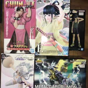 Row of colorful anime figure boxes on a wooden floor, featuring a pink-clad girl figure on the left and a blue-haired character center, plus a standing boy figure on the right and a metal dinosaur robot box at the bottom right.
