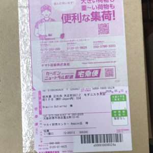 Pink Japanese delivery label on a cardboard box, showing barcode, QR code, and tracking text in Japanese.