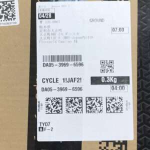 White shipping label stuck on a brown cardboard box, showing a barcode, multiple QR codes, and text like 'GROUND' and '0.3Kg' with a weight and time stamps (07:00, 04:00).