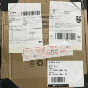 Cardboard parcel with multiple white shipping labels featuring barcodes, Japanese and Chinese text, and red stamps; black tape on edges.
