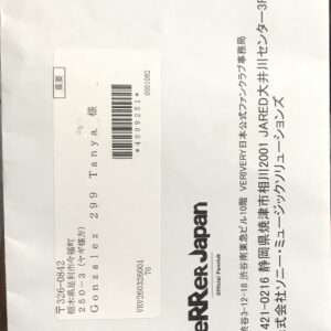 White mailing envelope with a postage label showing a barcode, recipient name/address in Japanese, and vertical text along the right edge; a circular stamp near the top-left.
