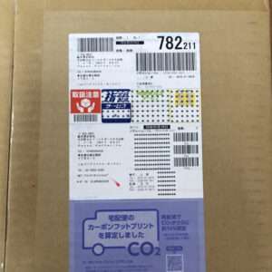 Cardboard box with a white shipping label featuring multiple barcodes and a red caution icon on the left side.