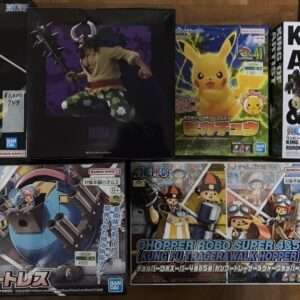 Assortment of Bandai Namco collectible figure boxes on a wooden floor, featuring anime characters (including a One Piece-style figure and a Pikachu box) arranged in two rows with a 'King of Artist' box on the right.
