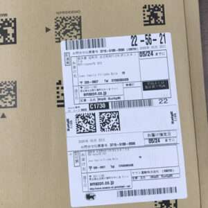Brown cardboard box with a white shipping label featuring multiple QR codes, barcodes, and Japanese text; label attached to the box’s surface.