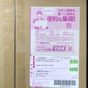 Pink parcel label on a brown cardboard box, featuring a delivery worker illustration, barcode, and Japanese text.