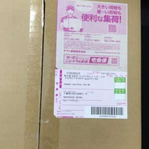 Cardboard shipping box with a large pink Japanese label showing recipient/address fields and a barcode text blocks.