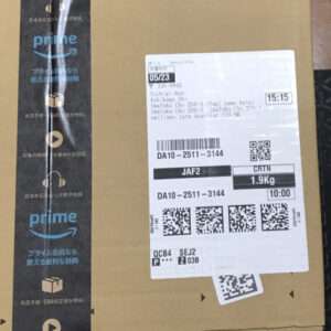 Brown Amazon package with Prime-branded tape on the sides and a white shipping label showing barcodes and QR codes on the front edge of the box, photographed indoors.