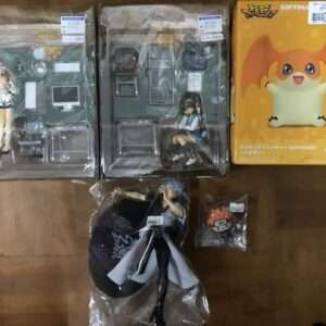 Assortment of anime figures and boxes on a wooden floor: two boxed figures in clear cases, a yellow SofviMates Digimon box, and a wrapped figure with accessories plus a small character head in a bag.