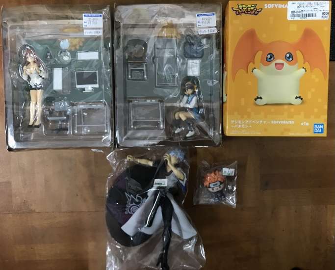 Screenshot - Articulos Japan (post Box Japan) Assortment of anime figures and boxes on a wooden floor: two boxed figures in clear cases, a yellow SofviMates Digimon box, and a wrapped figure with accessories plus a small character head in a bag. Articulos Japan (Post Box Japan)