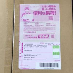 Cardboard box with a pink Japanese shipping label attached, showing a delivery form, a barcode, and a courier illustration on the label.