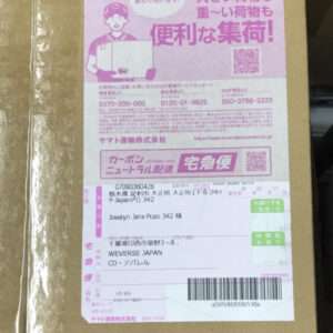 Pink Japanese delivery label on a brown cardboard box, showing a cartoon courier, large Japanese text, a QR code, and a barcode area at the bottom.