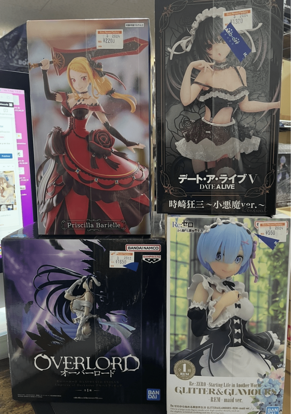 Ver Fotos Recientes 104 - Articulos Japan (post Box Japan) Shelf of boxed anime figures: left box features Priscilla Barielle in a red dress with a large sword-shaped weapon; right box shows a Fecha A Live figure in a black lace maid outfit with a choker; bottom left displays an Overlord figure with dark tones; bottom right shows Rem from Re:Zero in a maid outfit with blue hair. Articulos Japan (Post Box Japan)