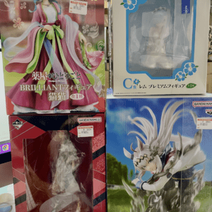 Stack of four anime figure boxes on a shelf: top-left pink-haired figure in a colorful dress, top-right white box with blue flower accents and a clear window, bottom-left red box with a figure visible through the window, bottom-right blue box featuring a dynamic white-haired figure.
