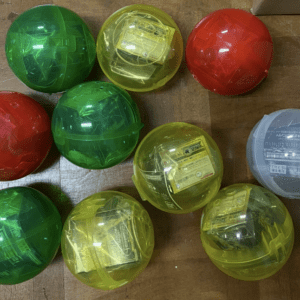 Top-down view of translucent toy capsules in red, green, yellow, and blue on a wooden floor grouped together with visible inner packaging inside some capsules.  Articulos Japan (Post Box Japan)