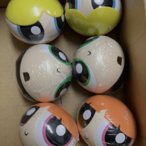 Six round, emoji-style balls with large cartoon eyes and bright hair colors (yellow, blue, green, orange) packed in a cardboard box.