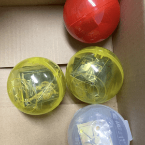 Box containing four round plastic capsules: red, two yellow translucent, and blue.