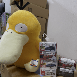 Large yellow plush toy with a pale beak and big circular eyes sits beside stacked boxes of toy cars.