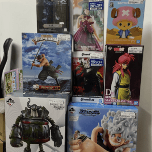 Assorted boxed anime figurines and collectibles stacked on a table, including action figures with swords, a pink-haired character, and a cartoon character in a hatbox.
