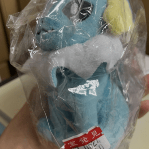 Blue plush toy dinosaur in clear plastic wrap, with a yellow ear and white belly, attached price tag (store item).