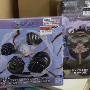 Two One Piece World Collectable Figures boxes on a shelf; left shows a fist-punching figure with energy rings, right shows a horned bearded character with a staff in purple packaging.