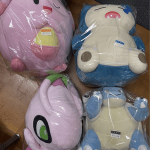 Four soft plush toys still in plastic packaging lying on a wooden floor: a pink round character with a big open mouth, a blue-and-cream creature with a red mouth, a lavender plush with a large green eye, and a blue-and-tan turtle-like plush far right.