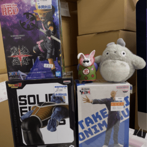 Stack of boxed anime figures on a shelf, including One Piece Red and Dragon Ball boxes, with a gray plush toy and a small bunny figurine nearby.