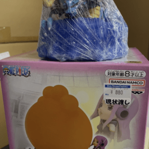 Pink box packaging for a One Piece toy with an orange spherical figure on the front; plastic bag with blue and yellow items placed on top.