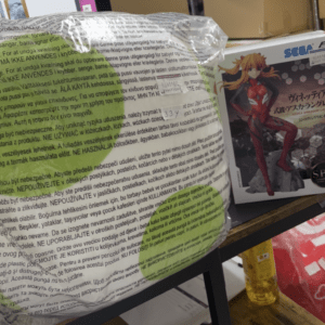 A large wrapped package covered in multilingual warning text sits on a shelf beside an anime figurine box.